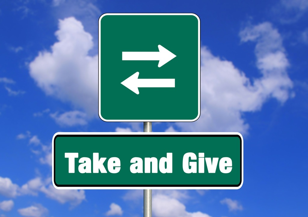 On a background of blue sky with fluffy white clouds are green street signs. One has arrows pointing in opposite directions and the other reads "take and give."