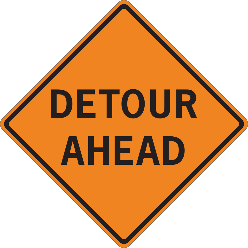 Square orange sign tilted so that one point of the square points upward, centered are the words "Detour Ahead"