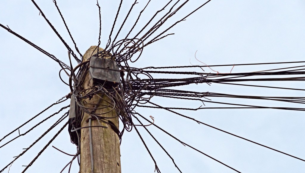 Top of a wooden pole is pictured with lines/wires coming from almost every direction in a tangled fashion.
