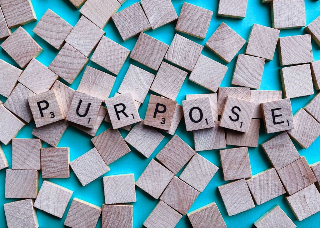 Blank wooden scrabble tiles spread randomly on teal colored background. Letter tiles spell out "purpose" mid-image horizontally. 