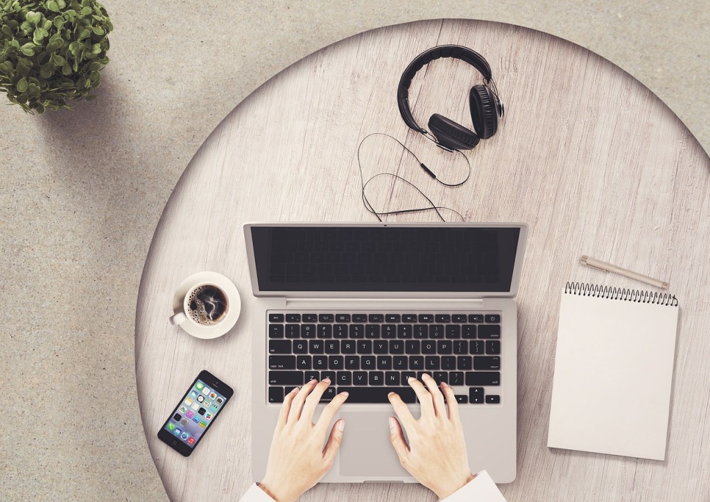 An overhead view of a woodgrain round table with a cell phone, a cup of coffee, a pair of headphones, a notepad and pen, along with a laptop with hands in the bottom center over the keyboard. 