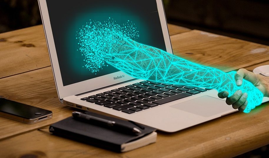 A digital arm is reaching out of a laptop computer to shake someone's hand. The laptop is sitting on a wooden top with a small note pad and pen next to the left side of the laptop and a cell phone directly above the note pad.